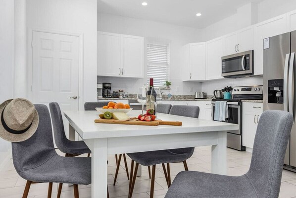A modern kitchen with white cabinets, a stainless steel refrigerator, and a dining area with grey chairs and a white table.
