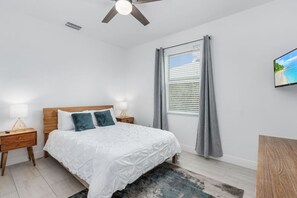 Un dormitorio con cama, mesitas de noche, ventilador de techo, ventana con cortinas y un cuadro en la pared.