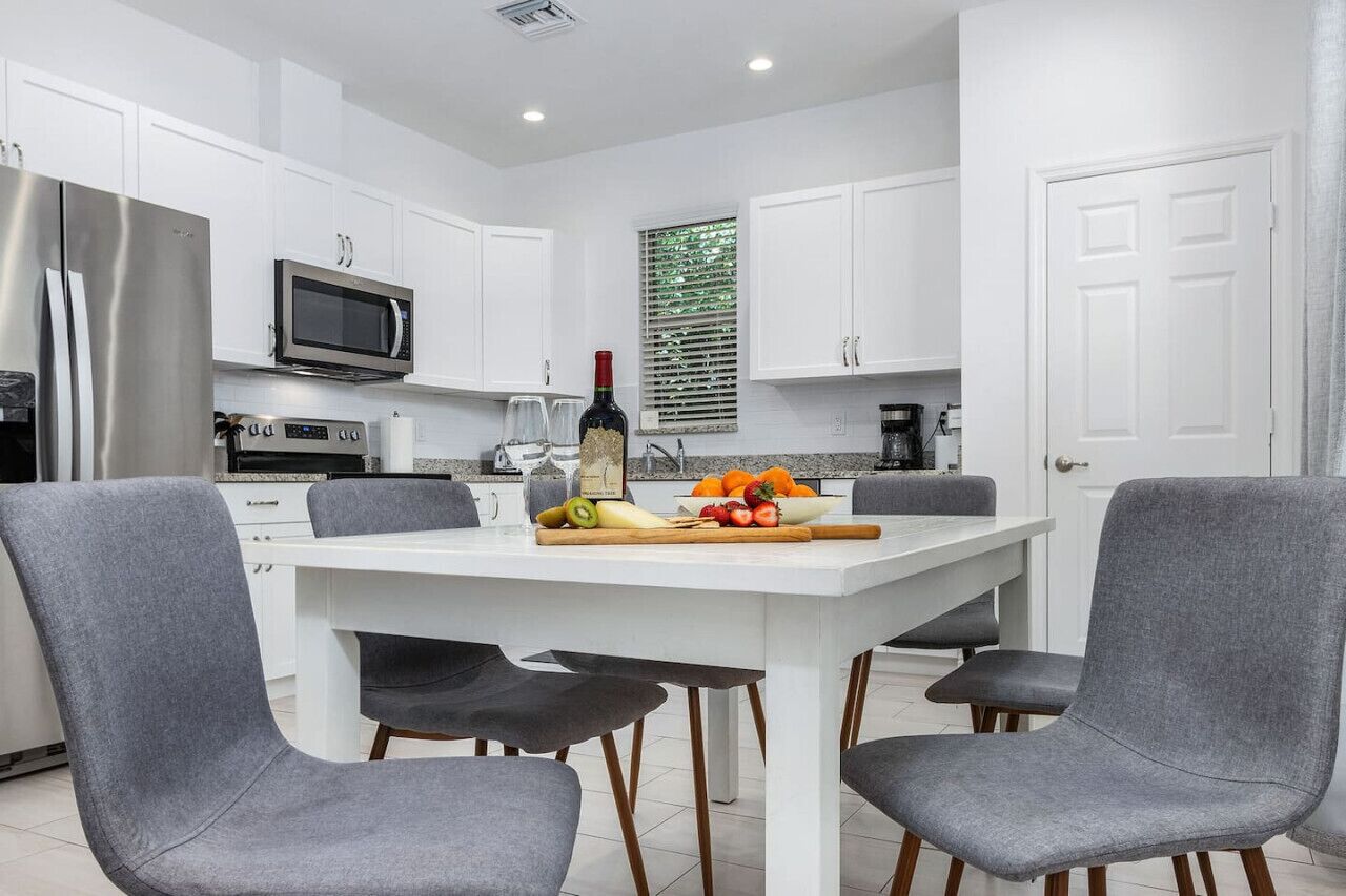 A modern kitchen with white cabinets, a stainless steel refrigerator, a microwave, and a dining table with four grey chairs.