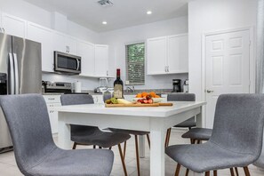 A modern kitchen with white cabinets, a stainless steel refrigerator, a microwave, and a dining table with four grey chairs.