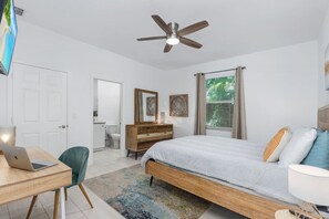 A bedroom with a bed, desk, chair, dresser, ceiling fan, and a window with blinds.