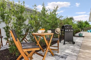 Un patio al aire libre con sillas de madera, una mesa redonda pequeña, una parrilla y plantas en macetas.