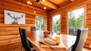 Een houten chalet van binnen met een eettafel, stoelen, een fruitschaal, wijnglazen en een brood.