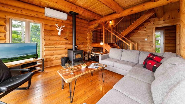 A cozy log cabin interior with a fireplace, a sofa, a television, and a wooden staircase. - Chalet Bois Rond 10: Hot Tub, Terrace & Views (Petite-Rivière-Saint-François)