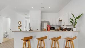 A modern kitchen with white cabinets, a stainless steel refrigerator, and a bar area with wooden stools. - Paradise at Hidden Palms Cove (Fort Myers)