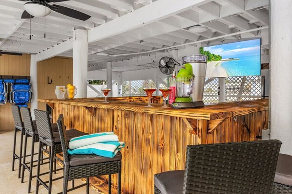A wooden bar area with bar stools, a television displaying a beach scene, and a ceiling fan. - Beach, pool time & fun times @ Blue Fish Lagoon (Fort Myers Beach)