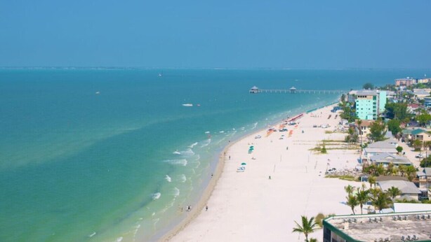 A beach with clear turquoise waters, a pier extending into the sea, and buildings with balconies overlooking the beach. - Beach, pool time & fun times @ Blue Fish Lagoon (Fort Myers Beach)