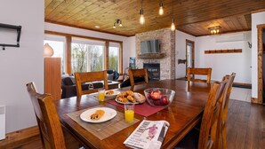 A dining area with a wooden table, chairs, breakfast items, and a newspaper. - 61B Chalet: Hot Tub, River Views & Wood Accents (Petite-Rivière-Saint-François)