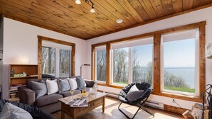 A cozy living room with a wooden ceiling, large windows, a sofa, a coffee table, and a chair. - 61B Chalet: Hot Tub, River Views & Wood Accents (Petite-Rivière-Saint-François)