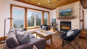 A cozy living room with a stone fireplace, large windows, and a comfortable seating area. - 61B Chalet: Hot Tub, River Views & Wood Accents (Petite-Rivière-Saint-François)