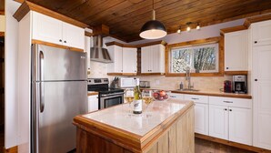 A modern kitchen with white cabinets, stainless steel appliances, a wooden island, and a large window with a view of trees. - 61B Chalet: Hot Tub, River Views & Wood Accents (Petite-Rivière-Saint-François)