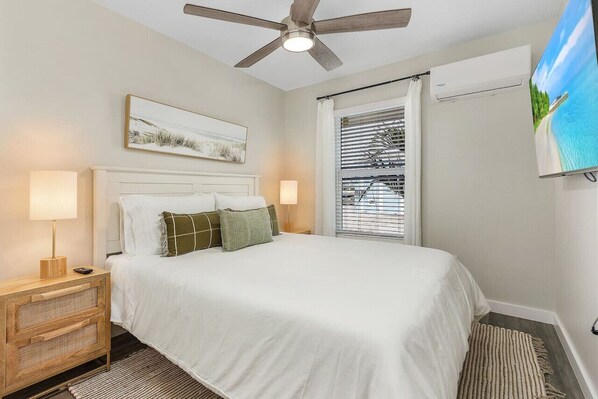 A bedroom with a bed, a ceiling fan, a window with blinds, a TV, and a nightstand with a lamp.