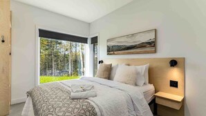 Une chambre moderne dotée d’une grande fenêtre, d’un lit avec une literie à motifs, d’une tête de lit intégrant un éclairage et d’un tableau représentant un paysage au-dessus du lit.