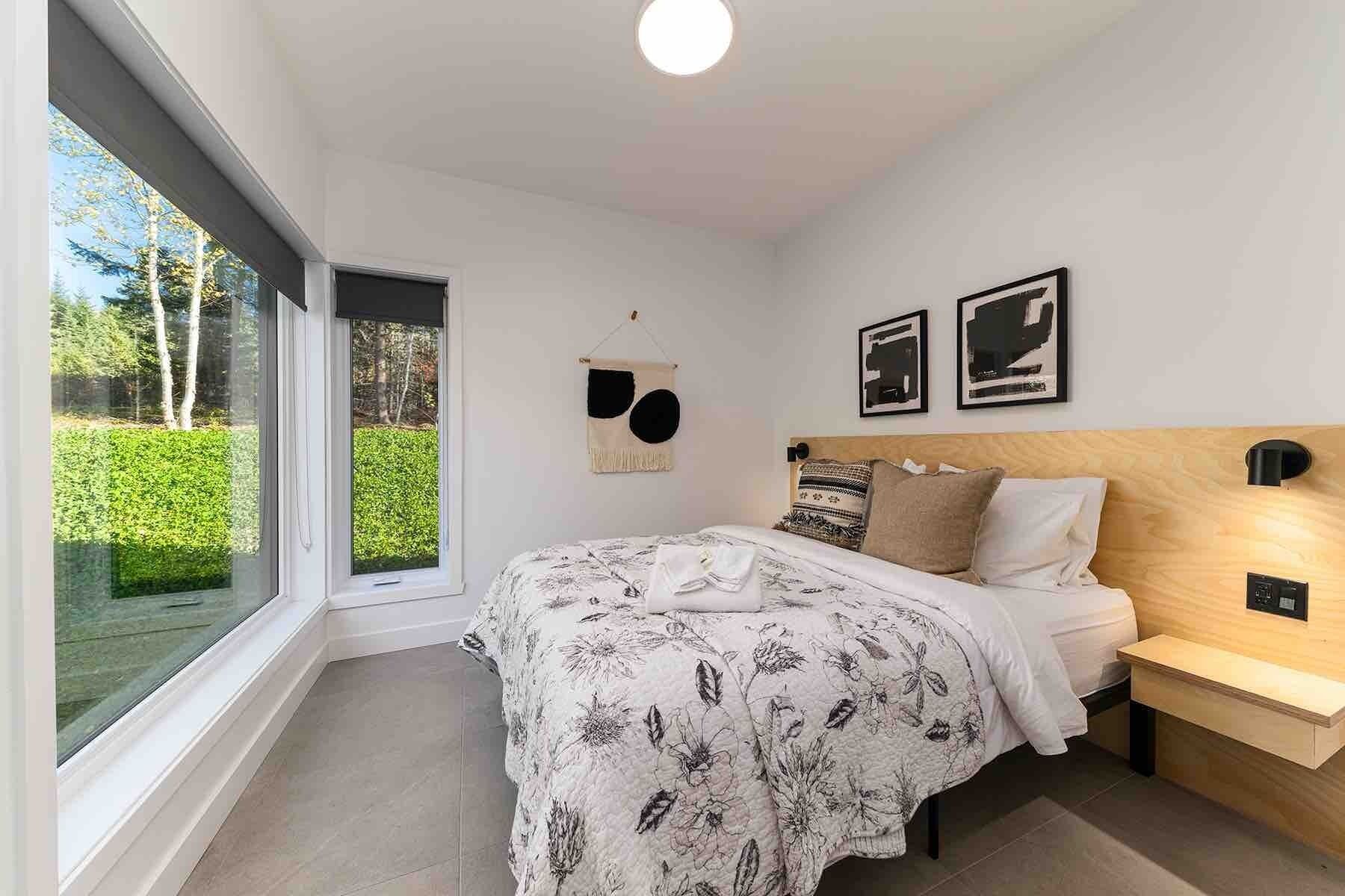 A modern bedroom with a large window, a bed with floral bedding, a bedside table, and two wall-mounted lights.