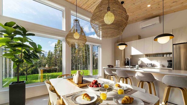 Une cuisine moderne équipée d’appareils électroménagers en acier inoxydable, un coin repas avec une table mise pour le petit-déjeuner, et de grandes fenêtres offrant une vue sur la verdure.