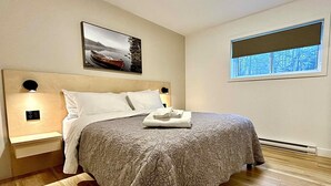 Una cama bien hecha con colcha estampada, dos lámparas de noche, una mesita de noche y una ventana con vistas a árboles.