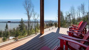 Une terrasse en bois avec des fauteuils Adirondack rouges, donnant sur une étendue d’eau et une zone boisée.