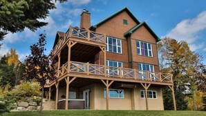 A two-story wooden house with a balcony, surrounded by trees and a grassy yard. - Condo 120B: Hot Tub, Panoramic Views & Game Area (Charlevoix)