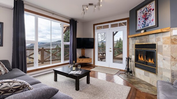 A spacious living room with a fireplace, a large TV, and a scenic view through the glass doors. - Condo 120B: Hot Tub, Panoramic Views & Game Area (Charlevoix)
