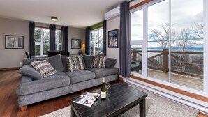 A modern living room with a grey sofa, a wooden coffee table, and large windows offering a view of a wooden deck and trees. - Condo 120B: Hot Tub, Panoramic Views & Game Area (Charlevoix)