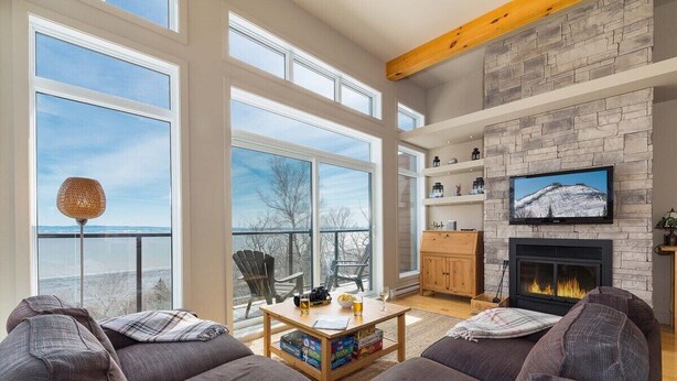 A spacious living room with large windows, a fireplace, and a television. - Le Méridien 33: Spa, River Views, Near Le Massif (Petite-Rivière-Saint-François)