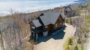 A large, two-story lodge with a stone foundation and a dark roof, situated in a forested area with a view of a lake and mountains in the background. - Le Méridien 33: Spa, River Views, Near Le Massif (Petite-Rivière-Saint-François)