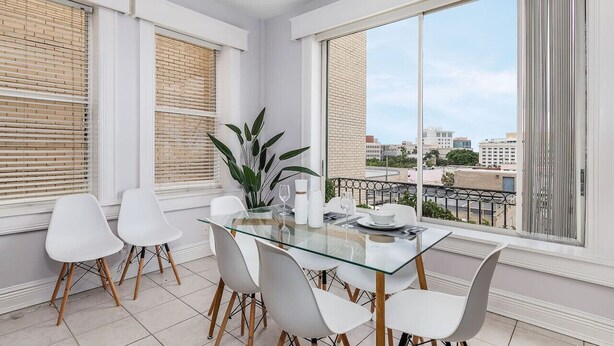 Un espace de repas moderne avec une table en verre et des chaises blanches, de grandes fenêtres équipées de stores et une vue sur le paysage urbain.