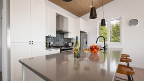 Een moderne keuken met witte kasten, een centraal eiland met barkrukken, een spoelbak en een raam met uitzicht op bomen.