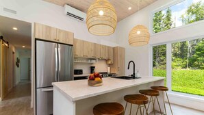 A modern kitchen with a large island, stainless steel appliances, wooden bar stools, and large windows offering a view of the outdoors. - La Charlebois 24: Forest Views, Hot Tub & Patio (Baie-Saint-Paul)