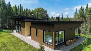 A modern house with large windows, a wooden exterior, and a gravel driveway. - La Charlebois 24: Forest Views, Hot Tub & Patio (Baie-Saint-Paul)