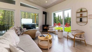 A spacious living room with a large sofa, a coffee table, and a fireplace. There are large windows offering a view of the outdoors. - La Charlebois 24: Forest Views, Hot Tub & Patio (Baie-Saint-Paul)