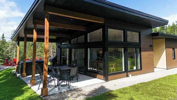 A modern house with a covered patio, large windows, and a well-maintained lawn. - La Charlebois 24: Forest Views, Hot Tub & Patio (Baie-Saint-Paul)