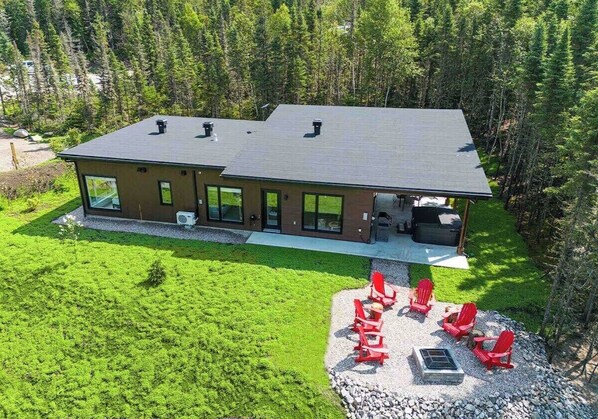 A modern two-story house with a flat roof, large windows, and a covered patio area with red chairs and a fire pit.