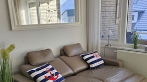 A modern living room with a sofa, a mirror, and a window with curtains. - Watt's Happiness Close to the Beach (Büsum)