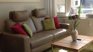 A modern living room with a brown leather sofa, green and red cushions, a glass coffee table, and a vase with flowers. - Watt's Happiness Close to the Beach (Büsum)