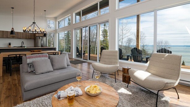A modern living room with a grey sofa, a round coffee table with snacks and drinks, and a view of the outdoors through large windows.