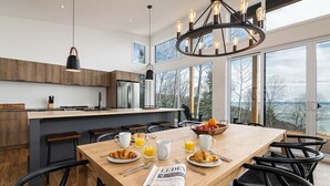 A modern kitchen with a wooden table set for breakfast, a chandelier, and large windows overlooking a scenic view.