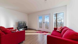 Ein modernes Wohnzimmer mit zwei roten Sofas, einer schwarzen Unterhaltungsanlage und einer Glastür, die auf einen Balkon mit Blick auf Bäume und Himmel führt.