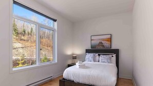 Ein Schlafzimmer mit großem Fenster, einem Bett mit weißer Bettwäsche, einem Nachttisch mit Lampe und einem gerahmten Bild an der Wand.