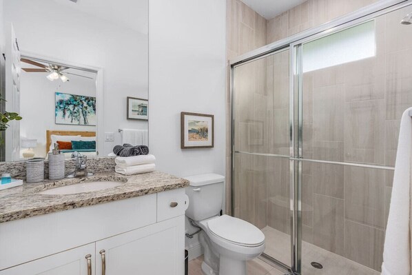 A modern bathroom with a granite countertop, white fixtures, a glass shower enclosure, and framed artwork on the walls.