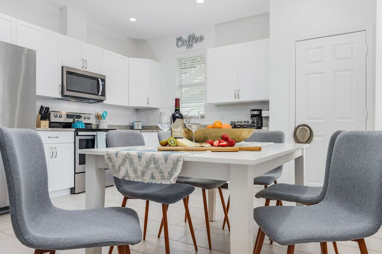 A modern kitchen with white cabinets, a stainless steel refrigerator, a microwave, and a coffee station. There are four grey chairs around a white table with fruits and a bottle.