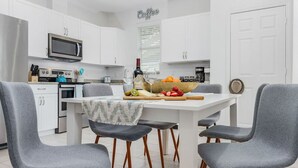 Une cuisine moderne avec des meubles blancs, un réfrigérateur en acier inoxydable, un four à micro-ondes et un coin café. Quatre chaises grises sont disposées autour d’une table blanche sur laquelle on trouve des fruits et une bouteille.