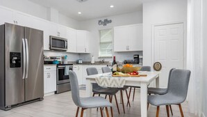 Une cuisine moderne avec des meubles blancs, des appareils en acier inoxydable et un coin repas avec des chaises grises et une table dressée pour deux personnes.