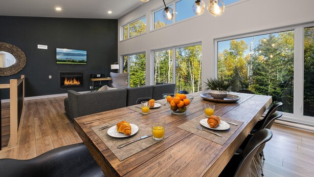 A modern dining area with a wooden table set for breakfast, large windows offering a view of trees, and a fireplace in the background. - Okanagan 97: Hot Tub, Pool Table & Summer Views (Les Éboulements)