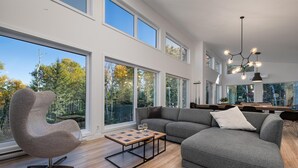 A modern living room with a grey sectional sofa, a unique chair, and a wooden coffee table. Large windows offer views of trees and a clear sky. - Okanagan 97: Hot Tub, Pool Table & Summer Views (Les  Éboulements)