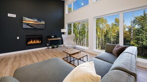 A modern living room with a fireplace, a large window with a forest view, and a cozy seating area. - Okanagan 97: Hot Tub, Pool Table & Summer Views (Les Éboulements)