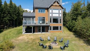 Une maison à deux étages dotée d’une grande terrasse et d’un foyer extérieur entouré de chaises.