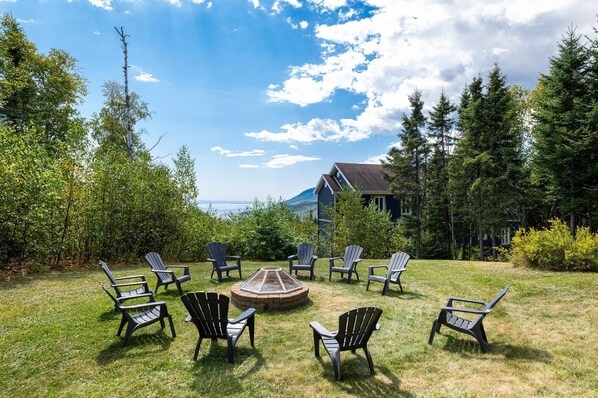 An outdoor seating area with Adirondack chairs arranged around a fire pit, surrounded by lush greenery and a view of a mountain and a house in the background. - Phoenix 75: Hot Tub, River Views & Pool Table (Charlevoix)