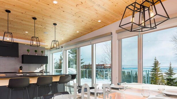 A modern kitchen with a wooden ceiling, large windows, and a dining area set for six. - Le Rupin 77: River, Mountain Views & Hot Tub (Charlevoix)