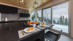 A modern kitchen with a large island, wooden ceiling, and large windows offering a view of trees and snow. - Le Rupin 77: River, Mountain Views & Hot Tub (Charlevoix)